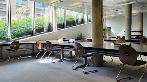 A section of the moore library glass room, showing desk spaces and large window panes, which follow a sloping line, getting larger as they descend toward the floor. The room is well lit by sunlight