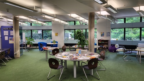 A section of the ground floor, showing large circular tables with games, jigsaws and books on them. In the background is a blue Acoustic Booth and a circular wall with a horizontal band of windows